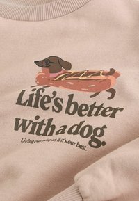 Light pink sweatshirt featuring a graphic of a brown dachshund in a hot dog bun, with the text "Life's better with a dog" in bold green.