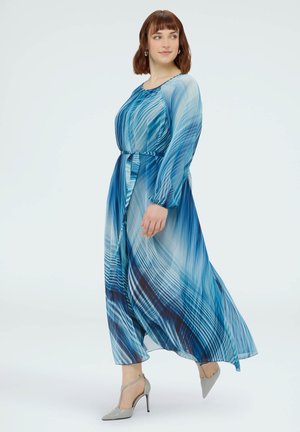 Žena s krátkými hnědými vlasy, oblečená v dlouhých modrých šatech s vzorem a šedými lodičkami na vysokém podpatku, kráčející a dívající se stranou na bílém pozadí.