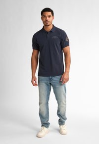 Námořnická polo košile s límcem a krátkými rukávy, s malým logem a nášivkou na levém rukávu, v kombinaci se světlomodrými džíny a bílými teniskami.