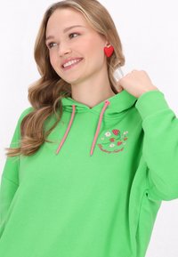 Felpa verde con lacci rosa, con fragole e fiori ricamati. La modella ha capelli ondulati e orecchini a forma di cuore rossi.