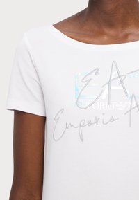 Camiseta blanca con escote amplio, mangas cortas y texto de logo bordado en plata sobre un fondo holográfico.