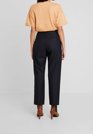 Frau trägt ein lockeres beiges T-Shirt, das in hoch taillierte schwarze Hosen und braune Stiefeletten mit Absatz gesteckt ist, und steht vor einem schlichten Hintergrund.