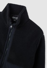 Giacca in pile nera con colletto alto, chiusura con zip e sezioni di tessuto liscio a contrasto. La texture è morbida e shaggy.
