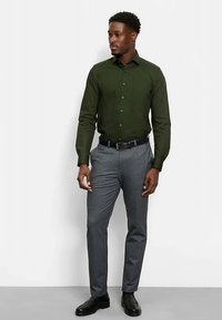 Dunkelgrünes Hemd mit Knöpfen und langen Ärmeln, kombiniert mit grauer, strukturierter Hose und schwarzem Gürtel, getragen mit polierten schwarzen Schuhen.