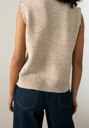 Beige gebreide mouwloze trui met een geribbelde onderrand, gecombineerd met donkere spijkerbroek. De stof ziet er zacht uit met een subtiele textuur.