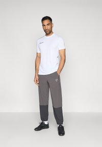 Λευκό αθλητικό μπλουζάκι κοντό μανίκι με μαύρες λεπτομέρειες, συνδυασμένο με γκρι παντελόνι με σχέδιο σε χρώματα και ελαστικές μανσέτες. Μαύρα αθλητικά παπούτσια.