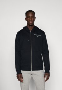 Schwarze Zip-Hoodie mit Kordelzugkapuze, Fronttaschen und weißem Logotext. Hergestellt aus einem weichen Stoff und mit gerippten Bündchen.