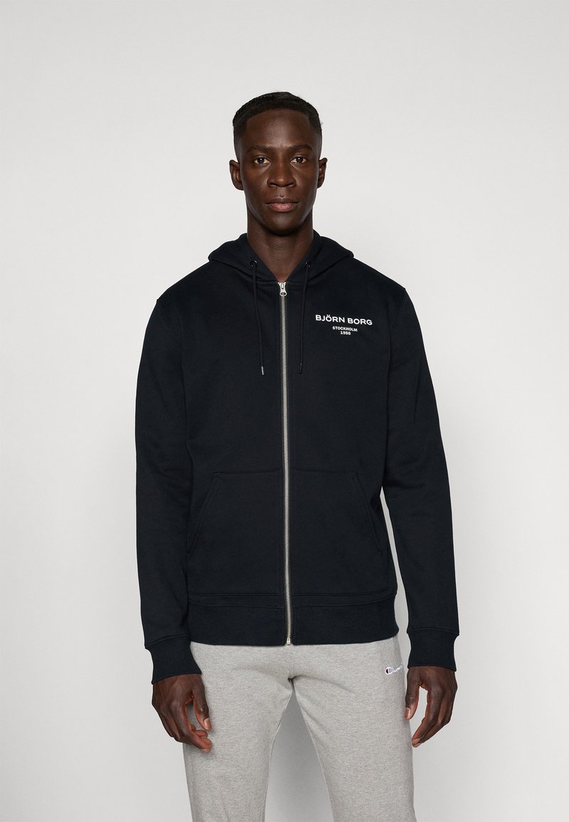 Schwarze Zip-Hoodie mit Kordelzugkapuze, Fronttaschen und weißem Logotext. Hergestellt aus einem weichen Stoff und mit gerippten Bündchen.
