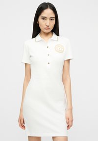 Νεαρή γυναίκα φορά ένα λευκό φόρεμα πόλο με κοντά μανίκια, χρυσά κουμπιά και χρυσό έμβλημα στο στήθος, στέκεται απέναντι σε ένα απλό φόντο.