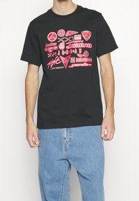 Camiseta de algodón negra con mangas cortas que presenta varios logotipos y diseños gráficos rojos centrados en la parte delantera, combinada con jeans de denim azul claro.