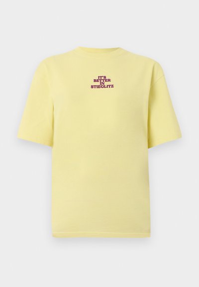 Κίτρινο κοντομάνικο μπλουζάκι με μοβ κείμενο κεντραρισμένο στο στήθος που γράφει «IT'S BETTER IN STIEGLITZ» σε ανοιχτό γκρι φόντο.