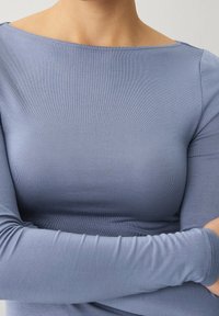 Pitkähihainen paita pehmeästä, teksturoidusta vaaleansinisestä kankaasta, leveällä venepäällä ja istuvalla siluetilla, joka esittelee sileää ompelua ja ei näkyvää metalliosaa.