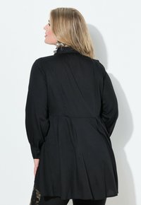 Schwarze Bluse mit langen Ärmeln, die eine fließende Silhouette, Knopfakzente an den Ärmeln, Faltendetails und eine glatte Textur aufweist.