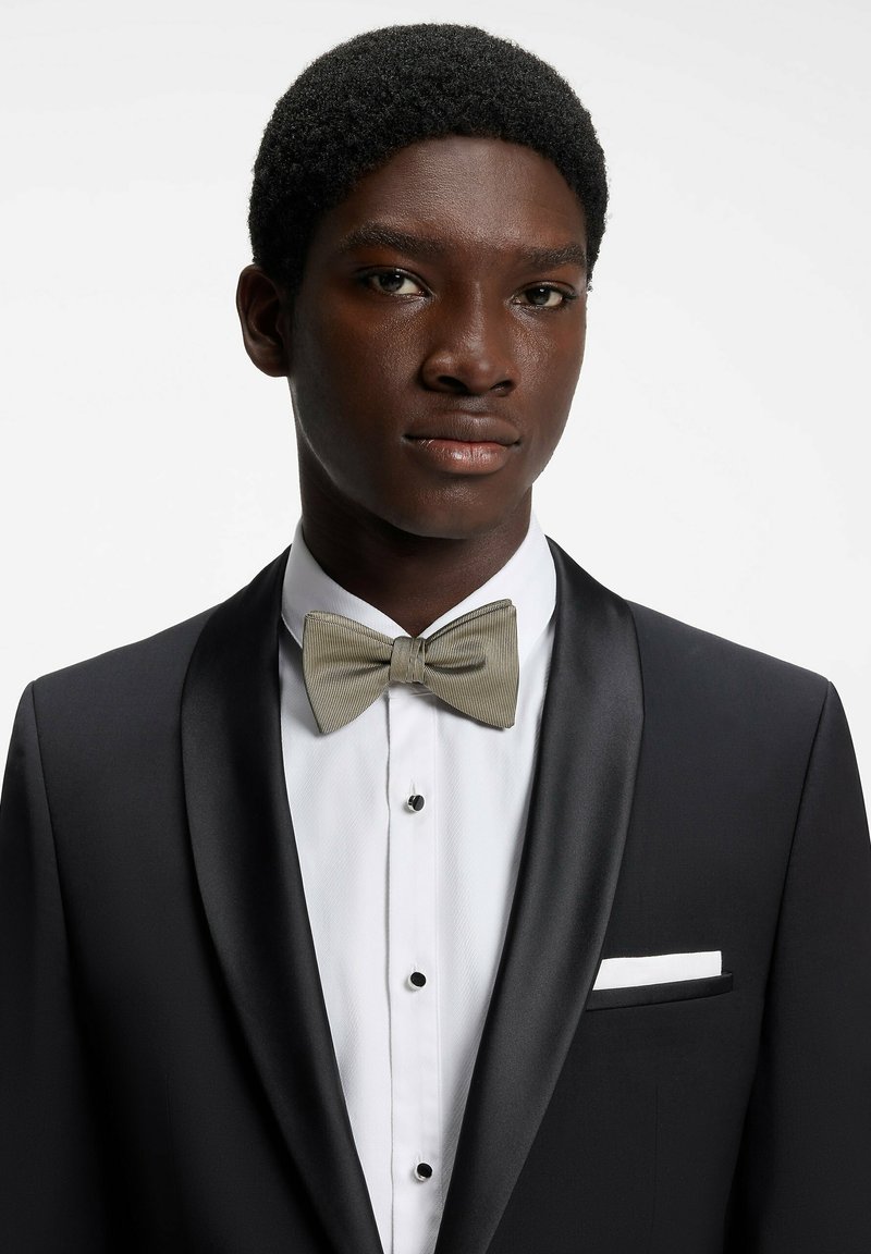 Jeune homme en smoking noir avec revers en satin, chemise blanche, nœud papillon beige et pochette blanche sur fond blanc uni.