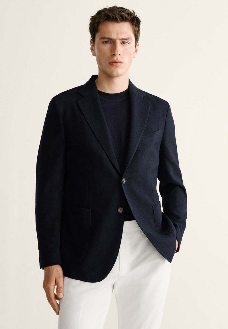 Joven con blazer azul marino sobre una camisa negra y pantalones blancos, de pie con una mano en el bolsillo contra un fondo liso.