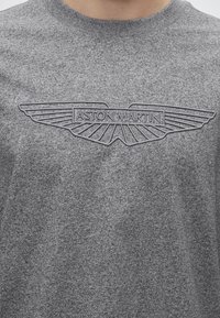 Γκρι πουλόβερ με κεντημένο λογότυπο της Aston Martin που διαθέτει στυλιζαρισμένα φτερά και το όνομα της μάρκας στο στήθος.