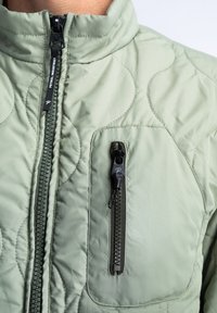 Hochkragenierte, hellgrüne Steppjacke mit Frontreißverschluss und einer Brusttasche mit kontrastierendem schwarzen Reißverschluss. Glatte Textur.