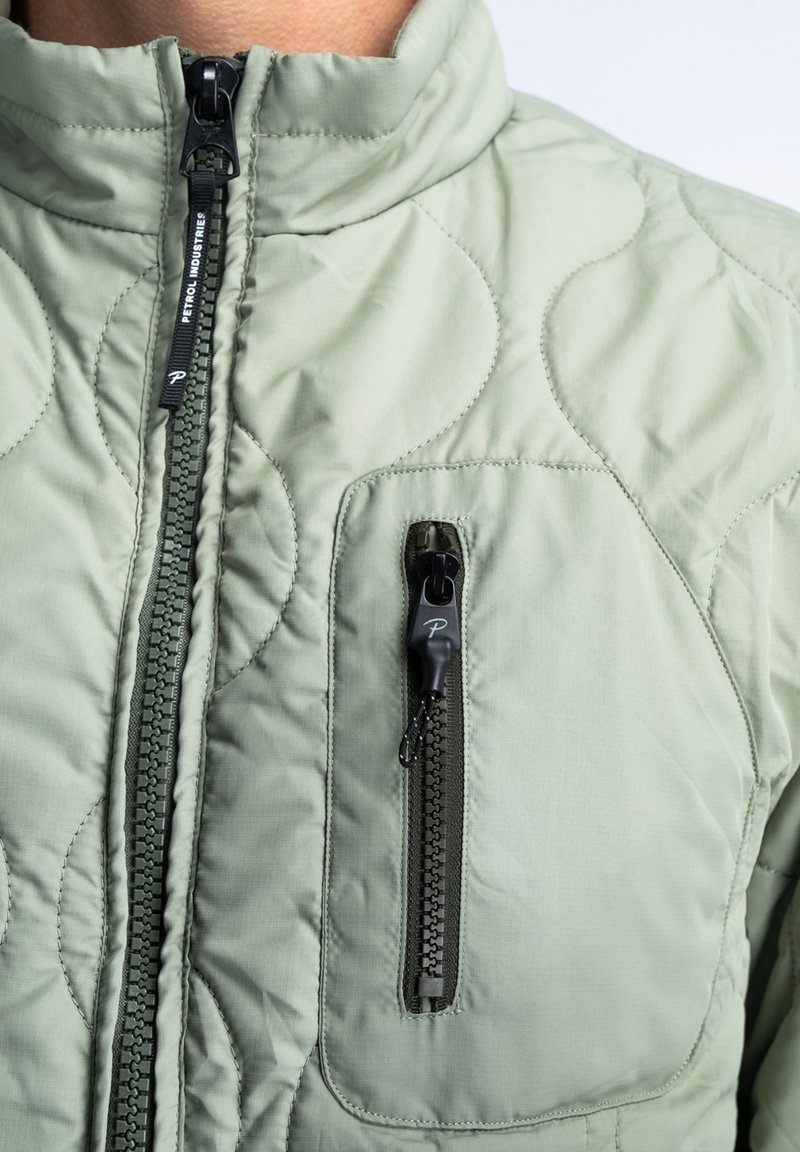 Chaqueta acolchada de color verde claro con cuello alto, que presenta un cierre frontal y un bolsillo en el pecho con una cremallera negra en contraste. Textura suave.