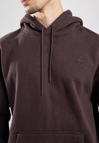 Felpa marrone in misto cotone, con tasca frontale, coulisse e una piccola patch con logo sul petto. Texture morbida.