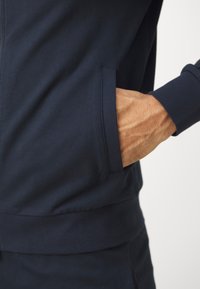 Felpa blu navy con vestibilità slim, dotata di tasca laterale e polsini a coste. Il tessuto sembra liscio con dettagli di cucitura minimi.