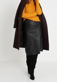Czarna skórzana ołówkowa spódnica o teksturowanym, dopasowanym kroju. Połączona z musztardowym swetrem z dzianiny i długim bordowym płaszczem. Czarne kozaki sięgające ponad kolano.