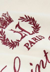 Fehér szövet, mély bordó hímzéssel. A design koronát, babérágakat és az "Párizs" szót elegáns betűtípussal tartalmaz.