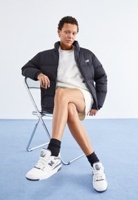 Svart dunjakke, hvit sweatshirt,beige skjørt og hvite joggesko med lilla aksenter. Stolen har en kromramme; bakgrunnen er lys blå.