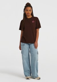 T-shirt en coton marron avec un petit logo graphique rose, associé à un jean cargo bleu clair à jambes larges, comportant de multiples poches et une coupe décontractée.