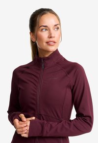 Bourgogne sportig zip-jacka med långa ärmar, slät textur, hög krage och sömnadsdetaljer. Logotyp synlig på framsidan.