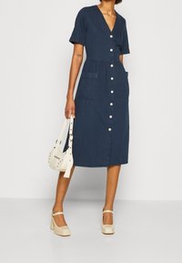 Kvinna i en marininfärgad klänning med knappar fram, som har fickor, bär en liten vit handväska och har på sig krämfärgade Mary Jane-skor.