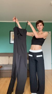 Schwarzer Jumpsuit mit verstellbaren Spaghetti-Trägern, tailliertem Oberteil und weit geschnittenen Hosenbeinen. Das Modell hält ihn vor einer grünen Wand.