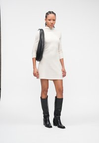 Rochie crem până la genunchi, cu model reiat și mâneci lungi, asortată cu cizme negre din piele până la genunchi și o geantă de umăr asortată. Design simplu, accente minimaliste.