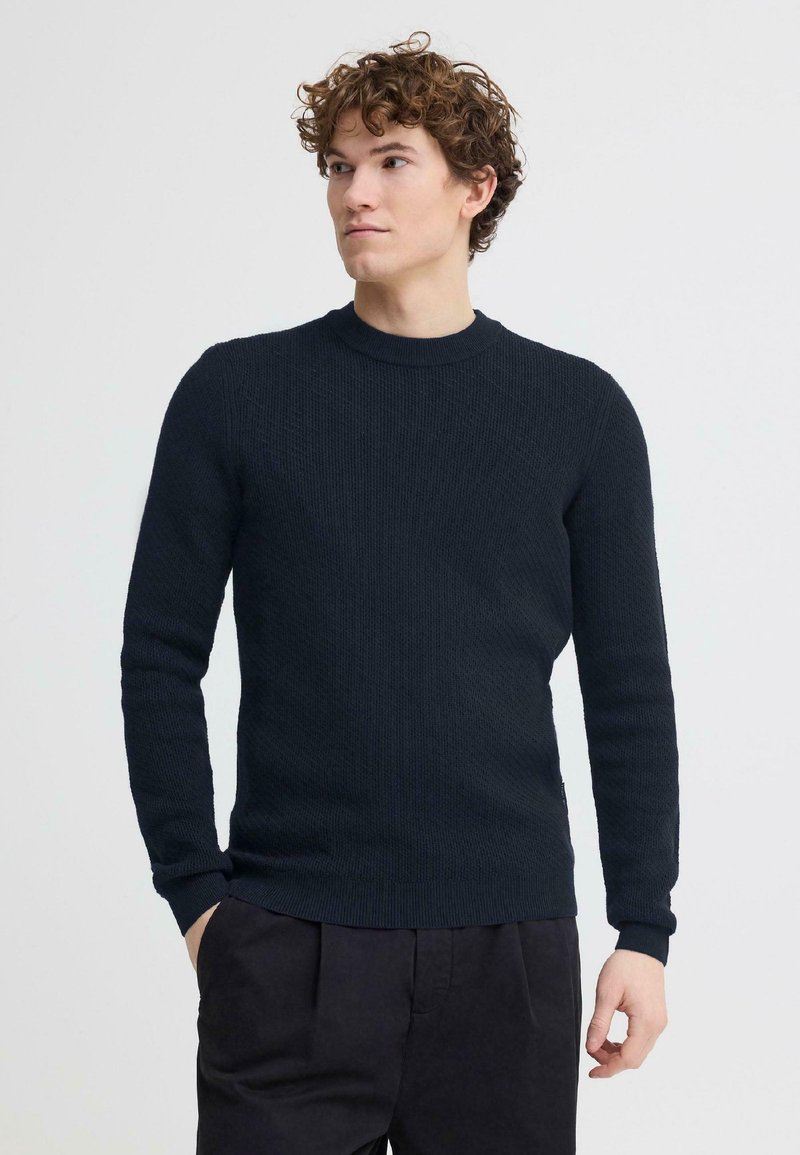 Jeune homme aux cheveux bouclés portant un pull ajusté bleu marine foncé et un pantalon noir, debout avec une main dans la poche, regardant sur le côté.