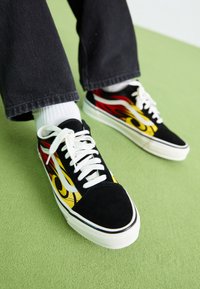 Zapatillas de ante negras con patrones de llamas amarillas y rojas, cordones blancos y suelas de goma, combinadas con calcetines blancos y pantalones vaqueros negros.