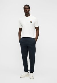 Άνδρας που φοράει λευκό μπλουζάκι με σχέδιο, στενά μπλε σκούρα παντελόνια και λευκά αθλητικά παπούτσια, στέκεται με τα χέρια στις τσέπες μπροστά σε απλό φόντο.