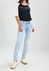 Casual outfit featuring a navy T-shirt with "TOMMY" text, white baseball sleeves, light blue jeans, and white sneakers with tan accents.