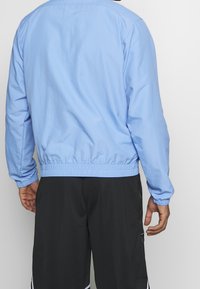 Ljusblå vindjacka med elastisk midja och ärmslut, med en slät yta och minimalistisk design. Svarta shorts fulländar looken.