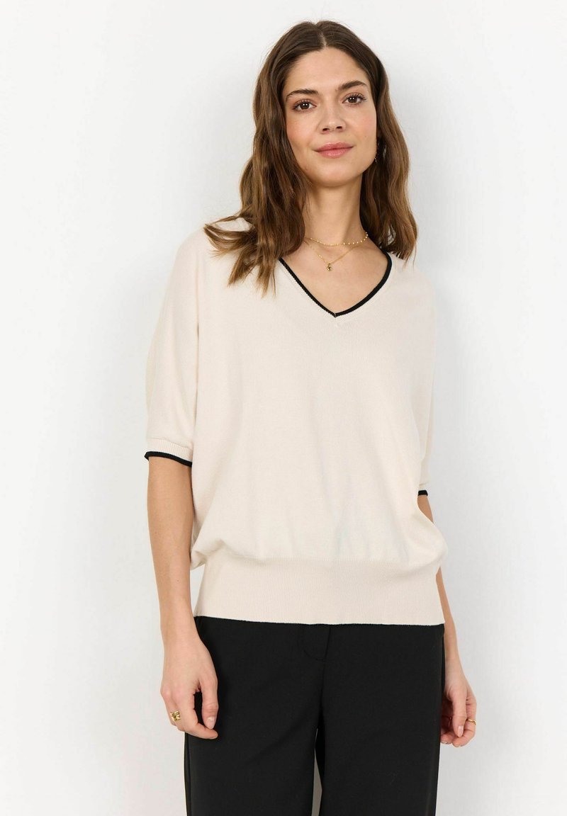 Cream-colored, short-sleeve knit top with a black trim along the V-neck and sleeves; loose fit with a ribbed hem. Paired with black trousers.