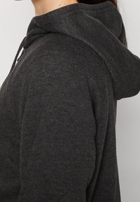 Sweat à capuche gris foncé avec un tissu texturé, des cordons au niveau du col, et un profil latéral montrant la capuche et la coupe décontractée.
