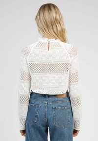 Haut en dentelle blanche à manches longues, motif structuré, col montant et détail en trou de serrure au dos. Associé à un jean bleu taille haute.