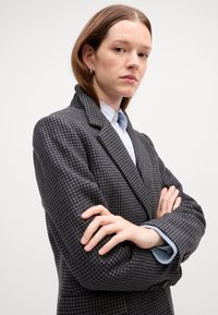 Blazer de espiga gris con solapas con muesca, tejido texturizado y dos botones. Puños de camisa azul claro visibles desde las mangas.