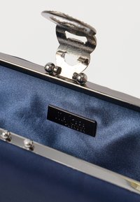 Ναυτικό σατέν εσωτερικό μιας τσάντας χειρός με μεταλλικό ασημένιο πλαίσιο και χαραγμένη πινακίδα επωνυμίας. Το κλείσιμο διαθέτει μια υφασμάτινη, διακοσμητική μπάλα.