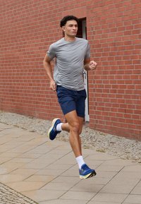 Un coureur portant un t-shirt de sport gris et un short bleu marine, avec des baskets bleues à semelles blanches. En arrière-plan, un mur en briques rouges.