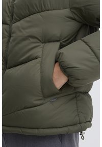 Olivovo zelená puffa bunda s horizontálne prešívanými sekciami, so zipsovým predným uzáverom, veľkými bočnými vreckami a nastaviteľným spodným lemom. Mäkká, zateplená tkanina.