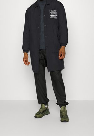 Svart lättviktig jacka med elastiska manschetter, prydd med flera vita "Lacoste"-logotyper. Burits över en blå tröja, i kombination med svarta byxor och gröna sneakers.