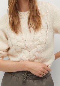 Pull en maille torsadée crème avec un motif texturé, col rond et manches courtes, associé à un pantalon de jogging gris.