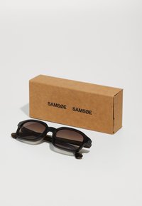 Occhiali da sole neri a forma quadrata con lenti gradienti marroni, dotati di montatura spessa e dettagli dorati sulle aste, accanto a una scatola di cartone marrone.