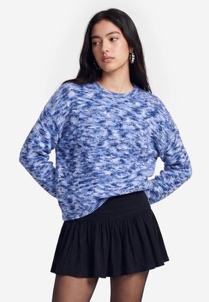 Jonge vrouw met lang donker haar, gekleed in een blauw gebreide trui met patroon, zwarte geplooide minirok en opvallende oorbellen, kijkt opzij.