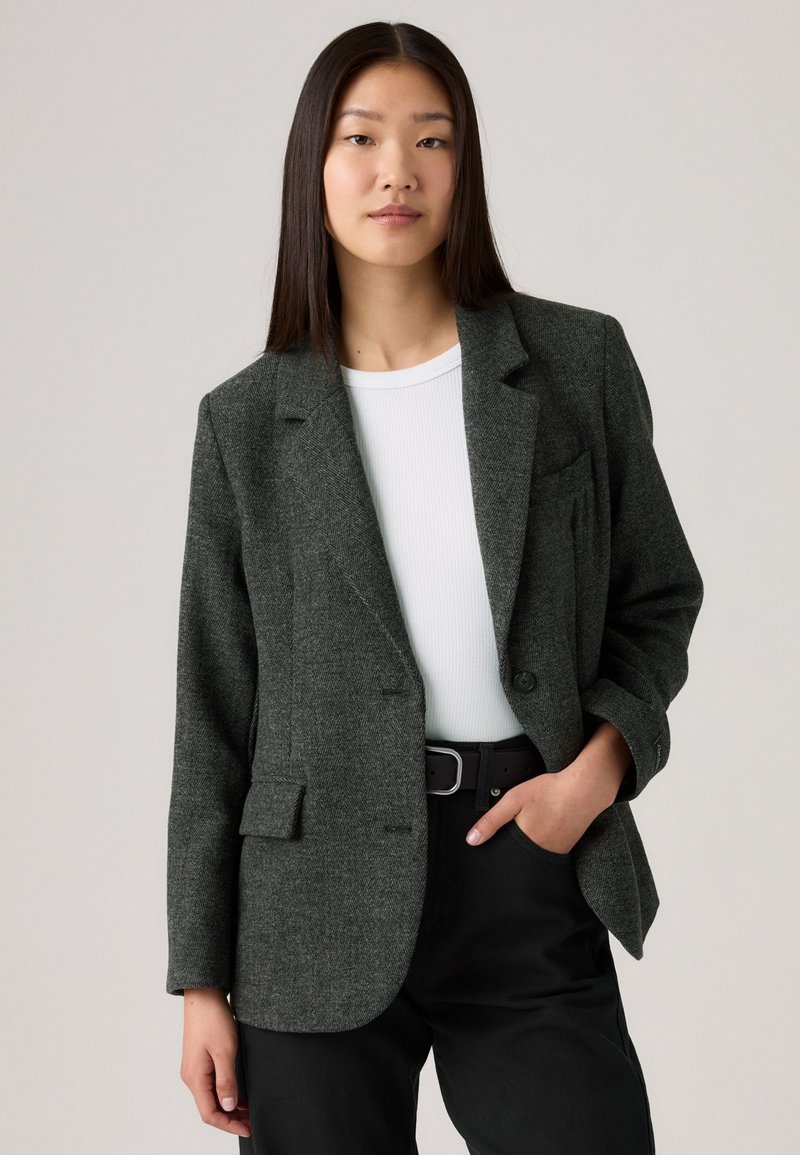 Grå tweedblazer med strukturerade axlar, framficka och enkel knappstängning, bärs över en vit topp och svarta byxor.