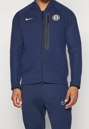 Mann trägt dunkelblauen Chelsea FC Trainingsanzug mit Nike-Logos, Reißverschlussjacke und passender Hose, steht vor schlichten Hintergrund.